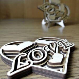 May include: A wooden heart-shaped ornament with the word "LOVE" layered and cut out in a decorative font. The ornament is made of multiple layers of wood, creating a 3D effect.