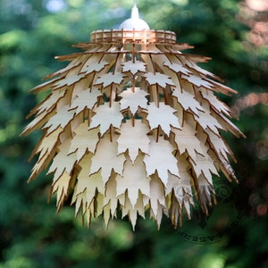 May include: A wooden pendant light fixture shaped like a sphere with a design of maple leaves. The leaves are cut out of the wood and create a delicate and airy look. The light fixture is hanging from a white cord.