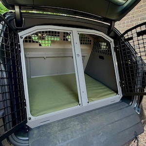 May include: A dual-compartment metal pet crate installed in the boot of a vehicle. The crate features a light grey frame, black wire mesh side doors, and two olive green padded floor mats. A black rubber protective mat extends from the base.