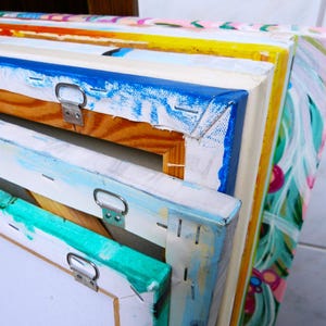 May include: A stack of colorful canvases with wooden frames and metal hanging hardware. The canvases are painted with abstract designs in various colors, including blue, green, yellow, and pink.