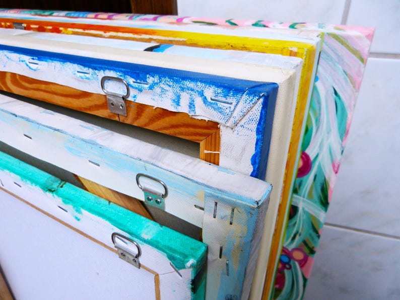 May include: A stack of colorful canvases with wooden frames and metal hanging hardware. The canvases are painted with abstract designs in various colors, including blue, green, yellow, and pink.