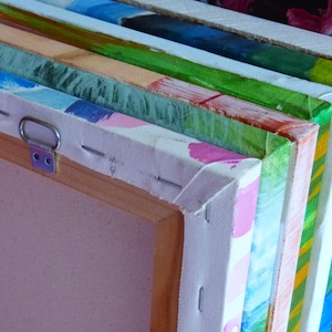 May include: Close-up of the edge of a stack of canvases, showing the wooden frames and the stretched canvas. The canvases are painted in various colors, including green, blue, pink, and yellow.