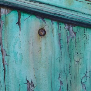 May include: Close-up of a weathered wooden surface painted in a teal green color. The paint is cracked and peeling, revealing the wood underneath. There is a rusty nail head in the center of the image.