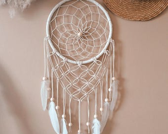 Puede incluir: Un atrapasueños circular con un patrón de red tejida en blanco dentro de un aro envuelto. El diseño incluye detalles de cordón de macramé, cuentas de madera clara y varias plumas blancas que cuelgan de la parte inferior.