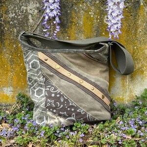 Könnte beinhalten: Olivgrüne Canvas-Umhängetasche mit Patchwork-Design. Die Tasche hat geometrische und florale Muster, akzentuiert mit beigen und braunen Streifen und goldfarbenen Nieten. Der verstellbare Riemen deutet auf ein lässiges Accessoire für den Alltag hin.