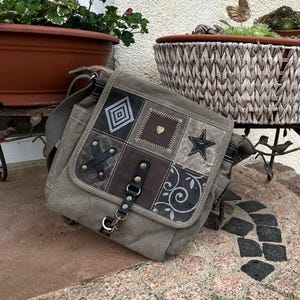 May include: A gray canvas messenger bag with a patchwork design on the flap. The patches include geometric shapes, a star, and floral patterns. The bag has a long adjustable strap and a metal clasp.