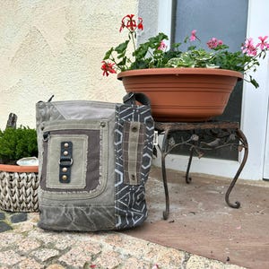Könnte beinhalten: Grau-, Oliv- und braunfarbene Canvas-Umhängetasche mit Patchwork-Design. Die Tasche hat eine Vordertasche mit schwarzem Lederakzent und Schnallenverschluss. Der Riemen ist auf der rechten Seite sichtbar. Die Tasche steht neben einem dekorativen Pflanzenständer. Auf der Seite der Tasche steht das Wort "Jumla".