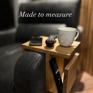 May include: A wooden side table with a built-in shelf for a remote control and a cup holder. The table is attached to the arm of a black leather sofa. The text "Made to measure" is visible in the image.