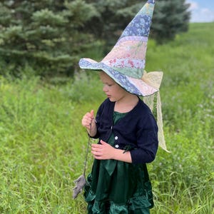 May include: A child wearing a whimsical witch costume outdoors. The costume includes a tall, pointed hat with a patchwork design in pastel colors, a dark blue cardigan, and a dark green dress with ruffled layers. The child is holding a gray toy.