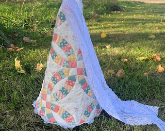 Könnte beinhalten: Eine handgefertigte, kegelförmige Steppdecke mit einem Patchwork-Design in Pastellfarben, darunter Rosa, Gelb und Blau. Ein weißer Spitzenschleier bedeckt die Steppdecke, die auf einer grasbewachsenen Oberfläche steht.