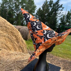 May include: A pointed witch hat featuring a patchwork design of orange and black fabric. The exterior displays patterns of crescent moons, stars, and broomsticks, while the underside of the wide brim is printed with a repeating orange pumpkin motif.