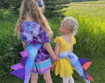 Könnte beinhalten: Zwei Kinder in einem grasbewachsenen Feld, gekleidet in Batik-Outfits und Drachenschwänzen. Eines trägt ein lila-blaues Batik-Oberteil und Shorts, das andere ein gelbes Kleid. Die Drachenschwänze sind blau, lila und orange.