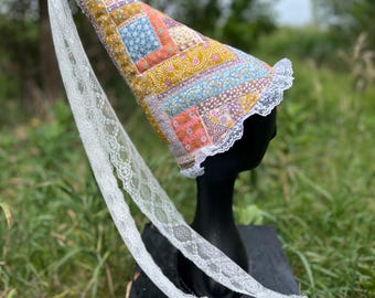 Könnte beinhalten: Ein kegelförmiger Hut mit einem Patchwork-Design aus Blumen- und geometrischen Mustern in Senfgelb, Orange und Blau. Der Hut ist am vorderen Rand mit weißer Spitze verziert und verfügt über einen langen, fließenden Schleier aus weißer Spitze.
