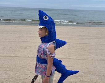 Könnte beinhalten: Ein Kind trägt ein blaues Hai-Kostüm an einem Sandstrand. Das Kostüm hat eine große Flosse und einen weißen Mund mit Zähnen.