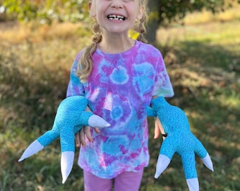Könnte beinhalten: Ein junges Mädchen trägt ein Batik-Shirt und eine lila Hose und hält zwei blaue und weiße Monsterkrallen. Sie lächelt und schaut in die Kamera.