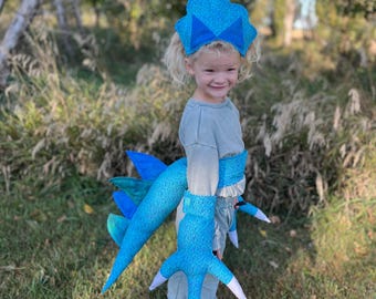 Könnte beinhalten: Ein Kinderkostüm mit einem blau-weiß gemusterten Kopfschmuck, Schwanz und krallenartigen Handschuhen. Das Kostüm hat ein skurriles Design mit einer Schleife auf dem Kopfschmuck und weißen Akzenten auf den Krallen. Das Kind trägt ein graues Hemd und eine Hose.