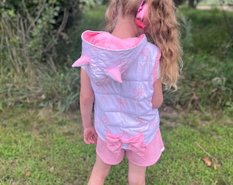 Könnte beinhalten: Ein Kind trägt eine rosa-weiße Weste mit Kapuze und passende Shorts. Die Weste hat rosa Akzente und eine Schleife auf dem Rücken. Die Shorts haben ebenfalls eine Schleife. Das Kind steht auf grünem Gras.