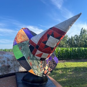 May include: A patchwork witch hat constructed from various fabric squares featuring orange, black, green, and purple colors. The design includes patterns of pumpkins, stars, and geometric shapes, with a pointed crown and a wide, curved brim.