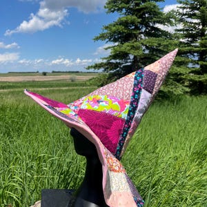 May include: A pointed hat constructed from a patchwork of various fabric patterns in shades of pink, purple, green, and black. The hat features a wide, slightly upturned brim and a tall, conical crown, all assembled with visible stitching.