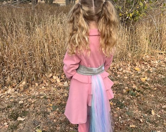 Könnte beinhalten: Ein junges Mädchen trägt ein rosa Einhornkostüm mit einem langen, bunten Schwanz. Das Kostüm besteht aus einem rosa Kleid mit einem Rüschen-Saum und einem passenden rosa Oberteil. Das Mädchen hat ihre Haare zu Zöpfen geflochten und steht auf einer braunen Wiese.