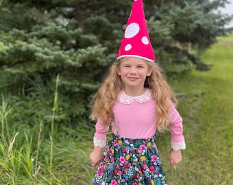 Könnte beinhalten: Ein Kind trägt ein rosa Kleid mit einem floralen Rock und einen roten Pilzhut mit weißen Punkten. Das Kleid hat einen weißen Spitzenkragen und Manschetten. Der Hintergrund zeigt grünes Gras und einen großen Nadelbaum.