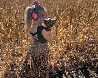 Könnte beinhalten: Ein junges Mädchen, das als Leopard verkleidet ist, trägt ein rosa Stirnband mit Katzenohren und einen rosa Kopfhörer mit Geräuschunterdrückung. Es steht in einem Feld mit getrockneten Maisstängeln. In der rechten Hand hält es einen Leoparden-Schwanz.