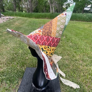 May include: A tall, pointed hat constructed from various fabric patches in shades of brown, orange, yellow, red, and green, featuring sunflower and leaf patterns. The hat has a wide, curved brim and is accented with a delicate, off-white lace ribbon.