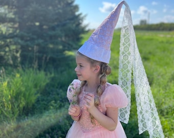 Könnte beinhalten: Ein rosa Prinzessinnenkleid mit Tüllrock und Pailletten-Oberteil. Das Outfit beinhaltet einen passenden rosa-weißen Hut mit Spitzenschleier. Das Mädchen hält einen kleinen Strauß lila Blumen.