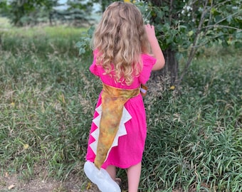 Könnte beinhalten: Ein junges Mädchen trägt ein rosa Kleid mit einem gelb-braunen Dinosaurierschwanz, der hinten befestigt ist. Das Mädchen steht in einem grasbewachsenen Gebiet mit Bäumen im Hintergrund.