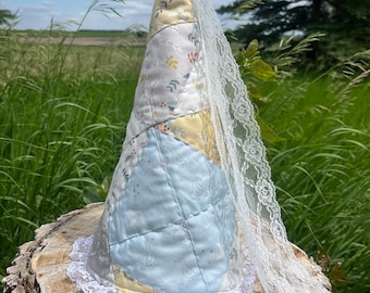 Könnte beinhalten: Ein kegelförmiger, gesteppter Stoffhut mit einem Patchwork-Design aus hellblauen, gelben und weißen Feldern mit dezenten Pflanzen- und Vogelmustern. Der Hut ist am unteren Rand mit weißer Spitze und einem langen Spitzenschleier verziert.