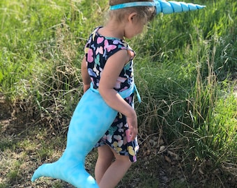 Könnte beinhalten: Ein kleines Kind verkleidet als Einhorn mit einem blauen Stirnband mit einem blauen und weißen Horn, einem blauen und weißen Schwanz und einem Blumenkleid.