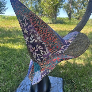 May include: A tall, pointed hat featuring a patchwork design of various batik fabrics in shades of orange, purple, teal, and black. The hat has a wide, curved brim and is constructed from multiple patterned textile segments with leaf and vine motifs.
