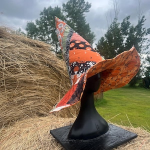 May include: A tall, pointed fabric hat featuring a patchwork design of various orange, black, and white Halloween-themed patterns, including pumpkins, stars, and moons. The wide brim is lined with an orange fabric printed with black pumpkin outlines.