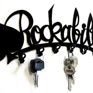 Könnte beinhalten: Schwarzer Metall-Schlüsselhalter mit dem Wort "Rockabilly" in dekorativer Schrift. Der Schlüsselhalter hat links ein Pik-Design und mehrere Haken für Schlüssel. Zwei Schlüsselsätze hängen an den Haken.