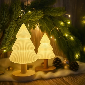 Op de afbeelding: Twee verlichte kerstboomvormige lampen met een warme gloed. De lampen zijn gemaakt van een geribbeld, gebroken wit materiaal en staan op houten voetstukken. De scène is versierd met dennentakken, lichtslingers en dennenappels.