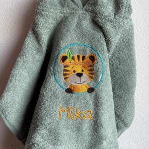 Puede incluir: Una toalla con capucha verde salvia con un rostro de tigre bordado en un círculo azul. El tigre es amarillo y naranja con una hoja verde. El nombre "Mika" está bordado en escritura amarilla debajo. La toalla está colgada.