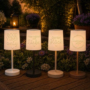 Könnte beinhalten: Vier Outdoor-Tischlampen mit weißen Schirmen und Sockeln. Jede Lampe hat eine andere Botschaft in weißer Schrift: "GOOD VIBES only", "Alles wächst mit Liebe", "Garten Glück" und "Lieblingsplatz". Die Lampen stehen auf einer Holzoberfläche.