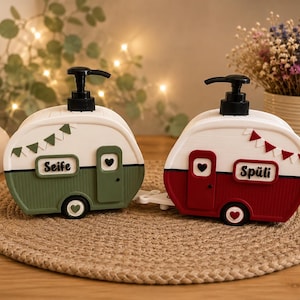 May include: Two caravan-shaped soap dispensers with black pump tops. One is green with the text Seife and a heart, the other is red with the text Spüli and a heart. Both feature white upper sections, decorative bunting, and heart-shaped accents on the doors and wheels.