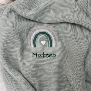 Op de afbeelding: Een saliegroene gebreide babydeken met een geborduurd regenboogontwerp in roze, groen en wit. De naam "Matteo" is eronder geborduurd. Een wit knuffelkonijn is zichtbaar op de achtergrond.
