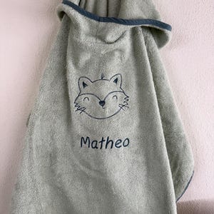 Peut inclure: Une serviette à capuche vert sauge avec une tête de renard brodée en gris foncé et le prénom Matheo cousu en fil gris foncé sous le renard. La serviette présente un liseré gris foncé le long des bords de la capuche et du corps principal.