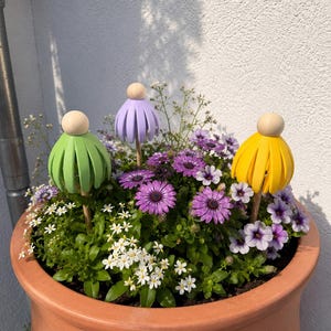 May include: A terracotta pot overflowing with a variety of flowers, including purple daisies and small white blossoms. Three decorative wooden flower toppers in green, purple, and yellow add a whimsical touch to the arrangement.
