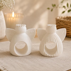 May include: Two white ceramic angel-shaped candle holders. The left angel has a smooth surface, while the right angel features vertical ribbed detailing. Both figures have circular openings in the centre, small wings, and a small heart embossed on each wing.