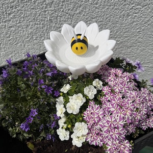 Peut inclure: Un abreuvoir blanc en forme de marguerite avec une figurine d'abeille jaune et noire au centre. L'abreuvoir est entouré d'une variété de fleurs colorées, dont des fleurs violettes et blanches, dans une jardinière noire.