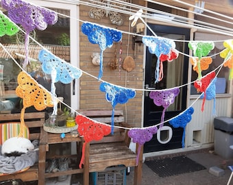 Könnte beinhalten: Eine mehrfarbige Häkelgirlande mit fächerförmigen Motiven in den Farben Lila, Grün, Blau, Orange und Rot. Jedes gehäkelte Teil ist an einer weißen Schnur befestigt und mit einer hängenden Quaste am unteren Ende versehen.