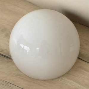 May include: A smooth, white, spherical object with a glossy finish. The round shape reflects the surrounding environment. The object is sitting on a light-colored wooden surface.