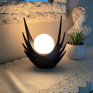 May include: A black ceramic table lamp with a white globe shade. The lamp has a unique design with two tall, pointed wings on either side of the globe. The lamp is sitting on a white lace table runner.