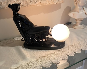 May include: A black ceramic lamp shaped like a reclining figure with a white globe shade. The lamp is on a white table with a lace trim.