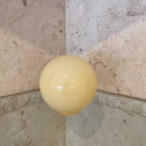 May include: A round, opaque, cream-colored glass globe. The globe is smooth and reflective, with a slight yellow tint. It sits on a clear, reflective surface, with a stone wall in the background.