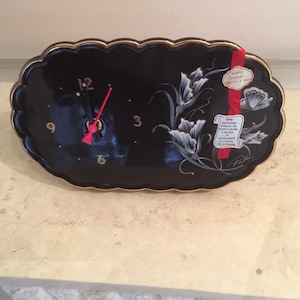 May include: Black oval clock with gold trim and a scalloped edge. The clock face has gold numerals and red hands. White floral design on the right side. A red ribbon with text is attached to the clock.