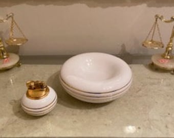 May include: A white ceramic ashtray and lighter set with gold trim. The ashtray is round with a recessed center. The lighter is spherical with a gold top. Two decorative gold scales flank the set.